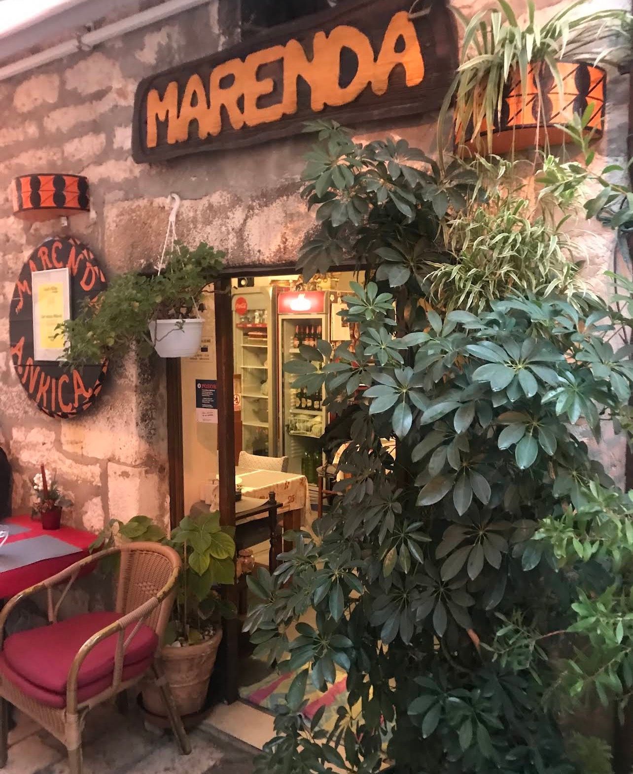 Ulaz u konobu Marenda 2: drveni natpis MARENDA na kamenom zidu, puno zelenih biljaka i pleteni stolac s crvenim jastukom uz vrata